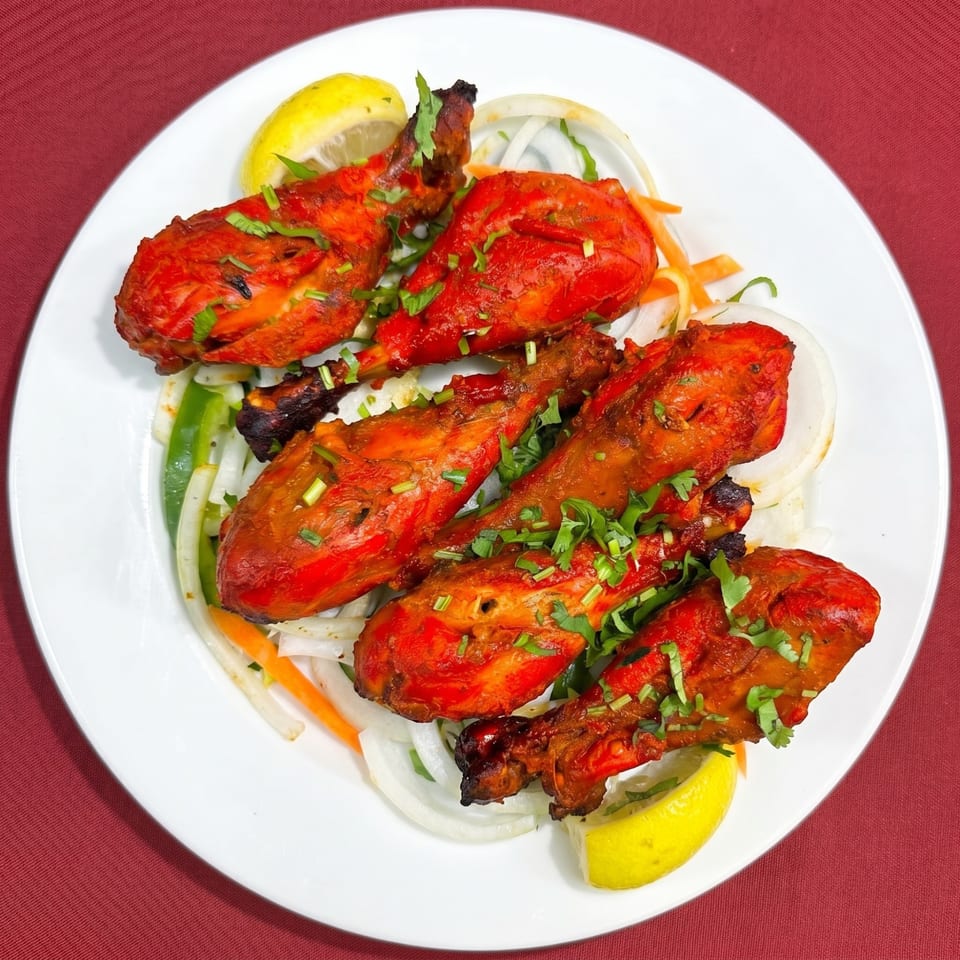 Six vibrant red tandoori chicken drumsticks, garnished with fresh herbs and lemon wedges, on a white plate.