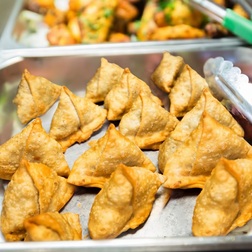 Golden-brown samosas, triangular fried pastries, are piled in a metal serving tray with tongs nearby.