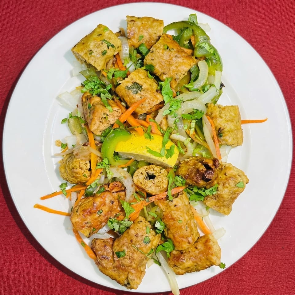 A white plate with golden-brown paneer tikka, green peppers, onions, carrots, cilantro, and a lemon wedge.
